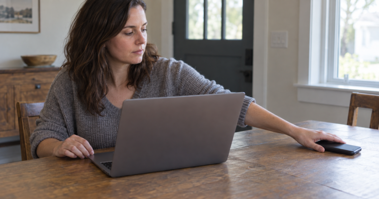 A woman at a laptop moves a face-down phone across the table.